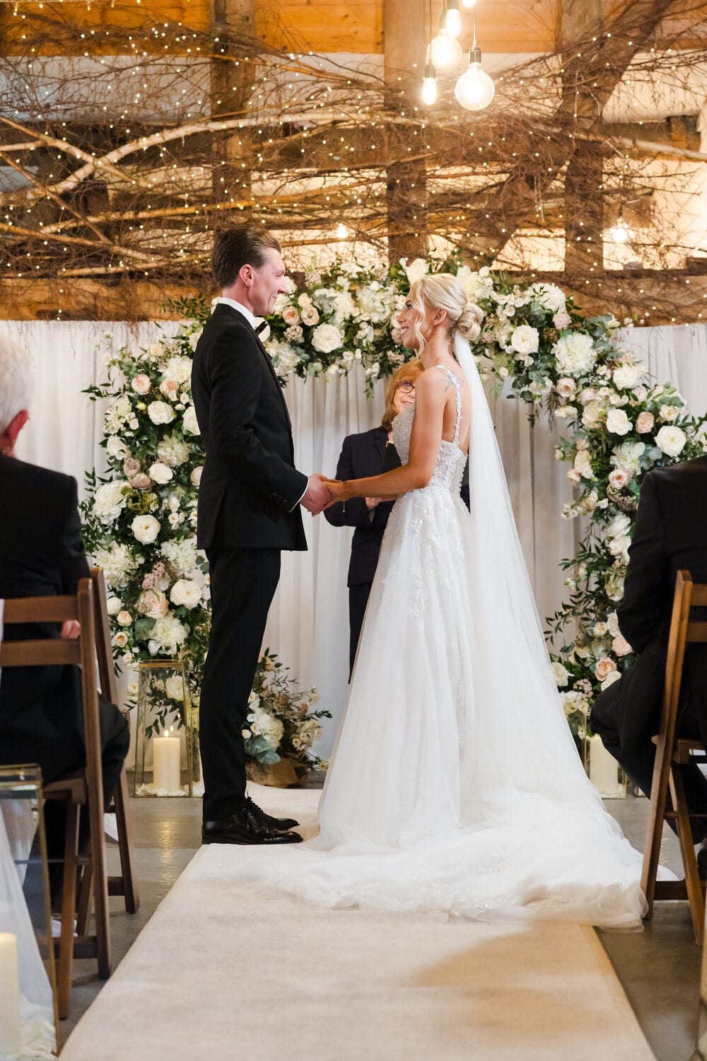 A couple exchange vows beneath a white floral arch in the Apple Barn