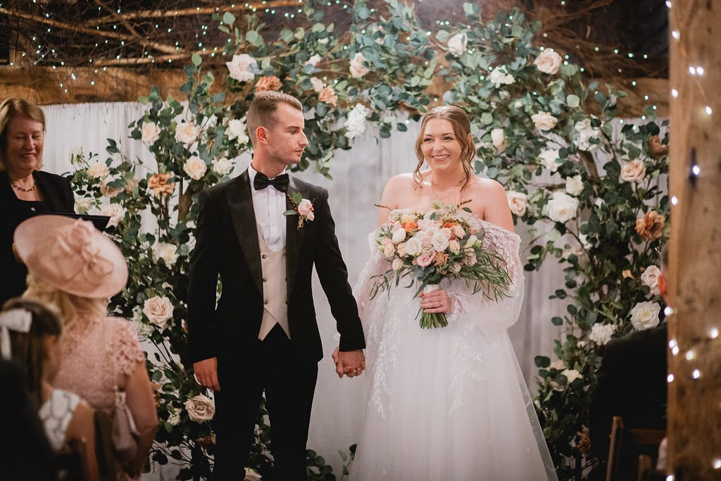 A couple beneath a white floral arch and fairy lights in the Apple Barn