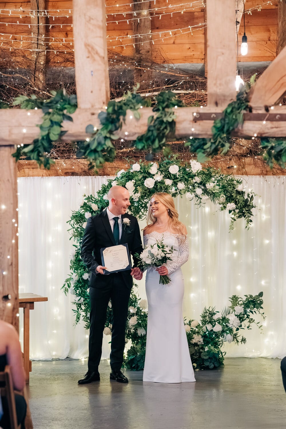 A couple and celebrant at a foliage arch in the Apple Barn